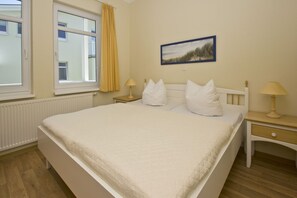 A bedroom with a bed, two bedside tables, a window with yellow curtains, a wall-mounted picture, and a radiator. - Balcony, free use of the AHOI adventure pool and sauna - Sea view apartment 46 - (Göhren)
