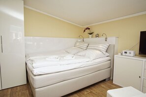 Una cama bien hecha con almohadas y toallas dobladas, una mesita de noche con una radio y una pequeña planta en maceta.