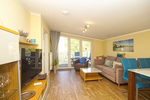 Ein gemütliches Wohnzimmer mit Sofa, Couchtisch und Essbereich mit Stühlen. Ein großes Glasfenster führt auf eine Terrassenfläche im Freien.