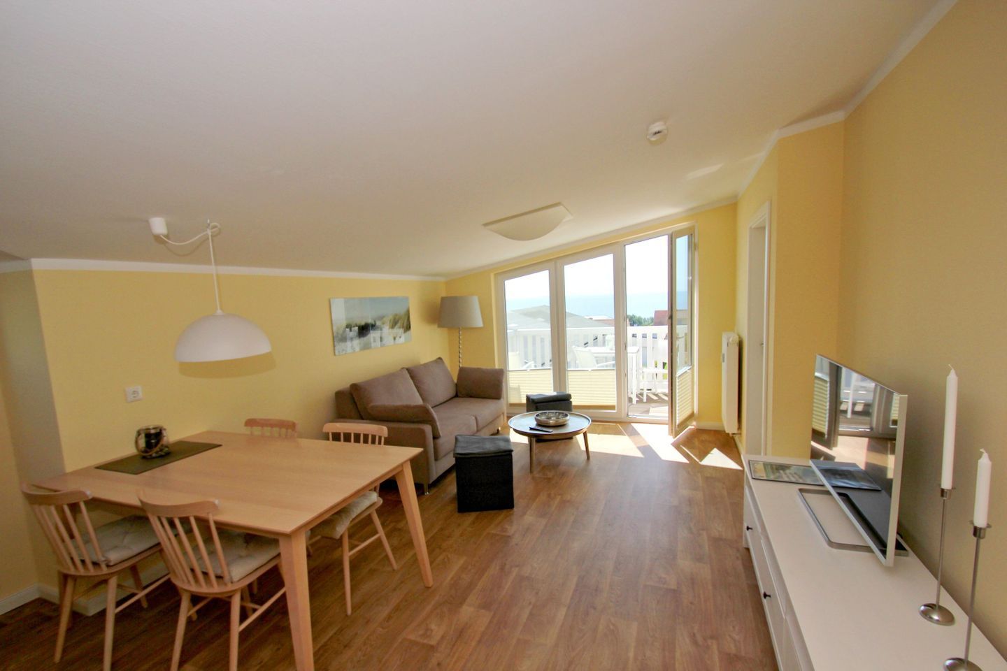 A spacious living area with a wooden dining table, two sofas, a TV, and a balcony with a view.