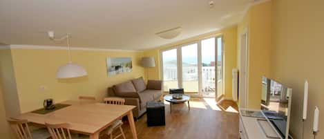 A spacious living area with a wooden dining table, two sofas, a TV, and a balcony with a view.