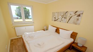 Un dormitorio con una cama de madera, ropa de cama blanca, una ventana con persianas, un radiador y una pared con tres cuadros florales.