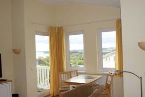 A room with a table and chairs, large windows with curtains, a white railing, and a view of greenery and buildings. - With a view of the sea and a balcony; access to the pool and sauna - Apartment 50 with a view of the sea - (Göhren)