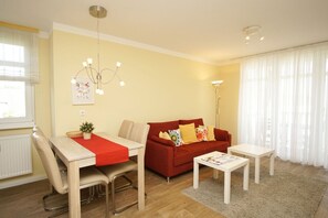 A modern living room with a red sofa, a dining table with chairs, a floor lamp, and a large window with sheer curtains. - Balcony, free use of the AHOI adventure pool and sauna - Apartment 56 with ocean view - (Göhren)