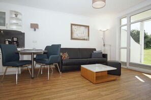 A modern living room with a black leather sofa, two blue chairs, a wooden coffee table, and a dining table with chairs. There is a painting on the wall and a floor lamp. - Terrace, free use of the AHOI adventure pool and sauna - Apartment 11 with ocean view - (Göhren)