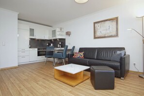 A modern living room with a black leather sofa, a wooden coffee table, and a dining area with blue chairs and a kitchen in the background. - Terrace, free use of the AHOI adventure pool and sauna - Apartment 11 with ocean view - (Göhren)