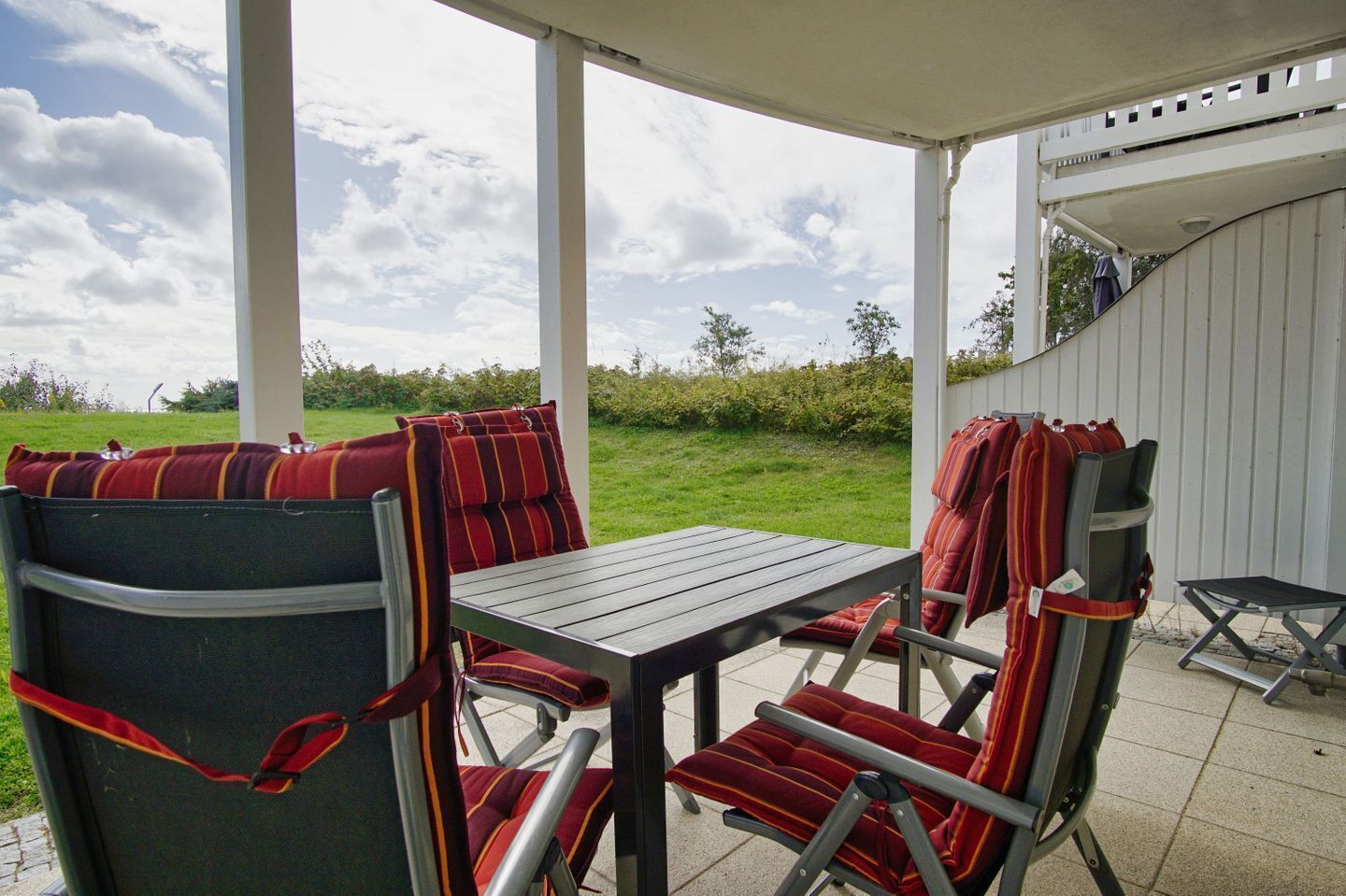 A covered patio with a table and chairs, a white wall, and a view of a grassy field.