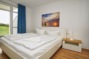 Ein modernes Hotelzimmer mit einem großen Bett, einem Nachttisch mit Lampe und einem Gemälde, das einen Sonnenuntergang über dem Wasser zeigt.