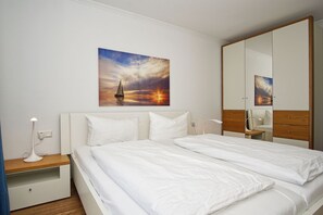 A hotel room with a bed, a nightstand, a bedside table, a lamp, a mirror, and a painting of a sunset over water. - With a view of the sea and a balcony; access to the pool and sauna - Sea View Apartment 14 - (Göhren)