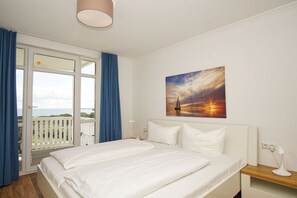 Une chambre  à coucher avec un lit, un tableau représentant un coucher de soleil, une fenêtre avec des rideaux et un balcon.