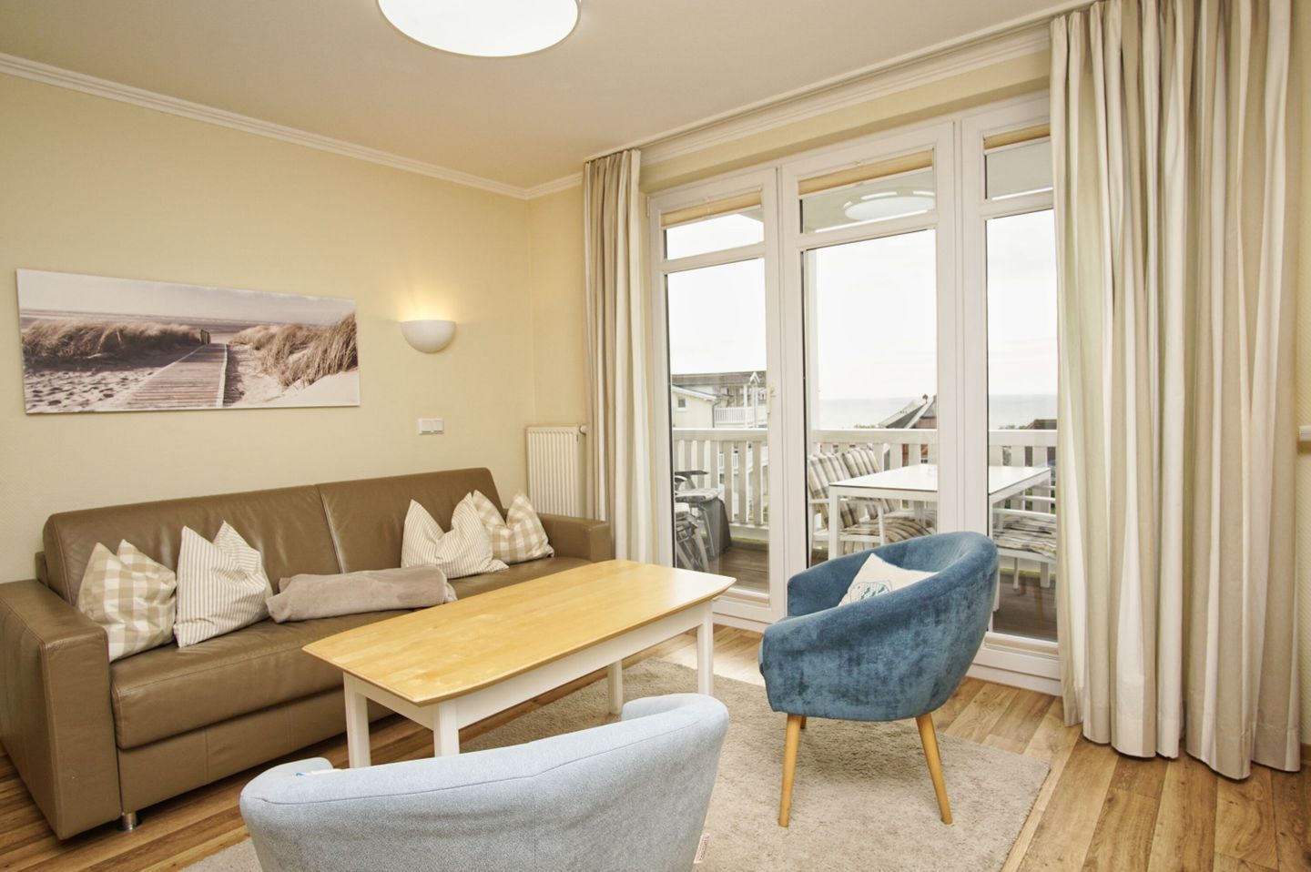 Un salon avec un canapé, une table basse et un fauteuil bleu. Il y a une grande fenêtre avec des rideaux, un tableau au mur et une vue sur la mer.