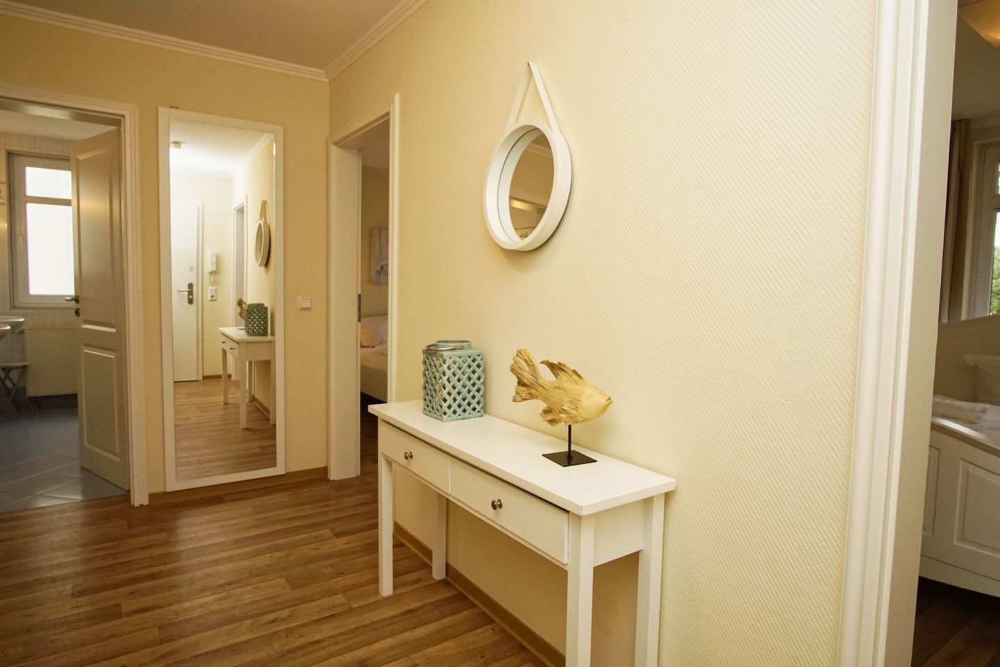 A hallway with a wooden floor, a white console table with a decorative fish sculpture, a round mirror, and a blue patterned container.