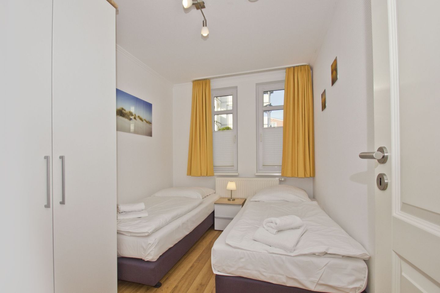 A bedroom with two beds, a nightstand, a window with yellow curtains, and a painting on the wall.