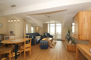 A spacious living room with a high ceiling, a dining area, and a kitchen. There is a large window providing natural light and a view of the outdoors. - With a view of the sea and a balcony, access to the pool and sauna - Sea View Apartment 27 - (Göhren)