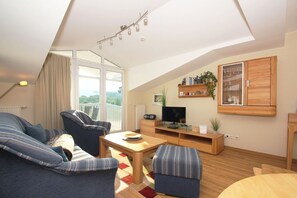 A modern living room with a sofa, armchair, and ottoman, a TV on a wooden stand, and a balcony with a view. - With a view of the sea and a balcony, access to the pool and sauna - Sea View Apartment 27 - (Göhren)
