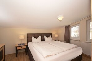 A hotel room with a large bed, two bedside tables, a nightstand, a window with curtains, and a ceiling light fixture. - With sea view and balcony, use of swimming pool and sauna - sea view apartment 27 - (Göhren)