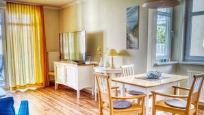 A room with a wooden floor, a white cabinet with a television, a dining table with chairs, and a window with curtains.