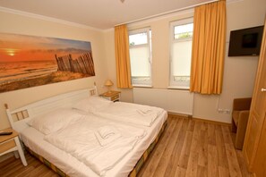 A bedroom with a bed, a nightstand, a lamp, a painting, a window with curtains, a radiator, and a television. - With a view of the sea and a balcony; access to the pool and sauna - Apartment 35 with a view of the sea - (Göhren)