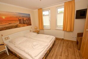 Ein Schlafzimmer mit Bett, Nachttisch, Lampe, Gemälde, einem Fenster mit Vorhängen, einer Heizung und einem Fernseher.