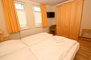 Ein Doppelbett mit weißer Bettwäsche, ein Holzschrank, ein an der Wand befestigter Fernseher und ein Sessel.