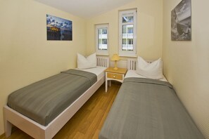 Ein kleines, sauberes Zimmer mit zwei Einzelbetten, einem Nachttisch mit Lampe und zwei Fenstern mit weißen Rahmen.