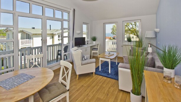 Un salón moderno con suelo de madera, muebles blancos, un televisor y ventanas grandes con vistas al mar.