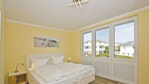A double bed with white bedding, a bedside table with a lamp, a large window with shutters, and a painting on the wall.