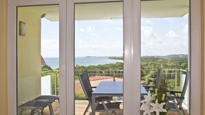 A balcony with a table and chairs, overlooking a coastal landscape with greenery and a view of the sea.