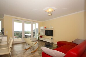 A modern living room with a red sofa, a rocking chair, a flat-screen TV, and a balcony with a view.