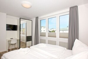A hotel room with a bed, a desk, a chair, a TV, and large windows with curtains. - With sea view and balcony, use of swimming pool and sauna - sea view apartment 64 - (Göhren)