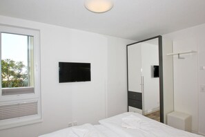 A modern hotel room with a large bed, a flat-screen TV mounted on the wall, a window with blinds, and a mirror cabinet.