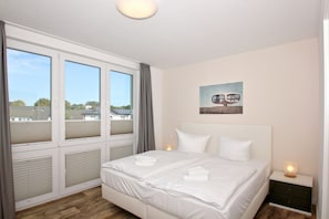 A bedroom with a large window, a bed with white linens, a nightstand with a lamp, and a wall-mounted picture. - With a view of the sea and a balcony, access to the pool and sauna - Sea View Apartment 64 - (Göhren)