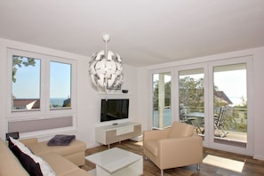 A modern living room with a flat-screen TV, a white console table, beige sofas, and a large glass door leading to an outdoor area. - With a view of the sea and a balcony, access to the pool and sauna - Sea View Apartment 64 - (Göhren)