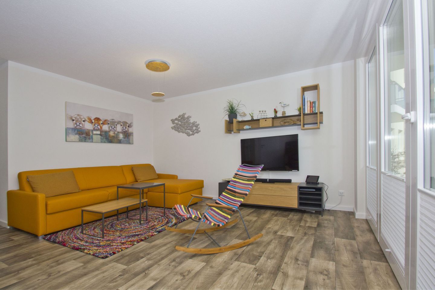 Ein modernes Wohnzimmer mit einem gelben Sofa, einem farbenfrohen Schaukelstuhl, einem Flachbildfernseher und Holzboden.