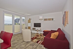 Ett modernt vardagsrum med en röd soffa, en vit fåtölj, en tv och ett stort fönster med utsikt över omgivningen.