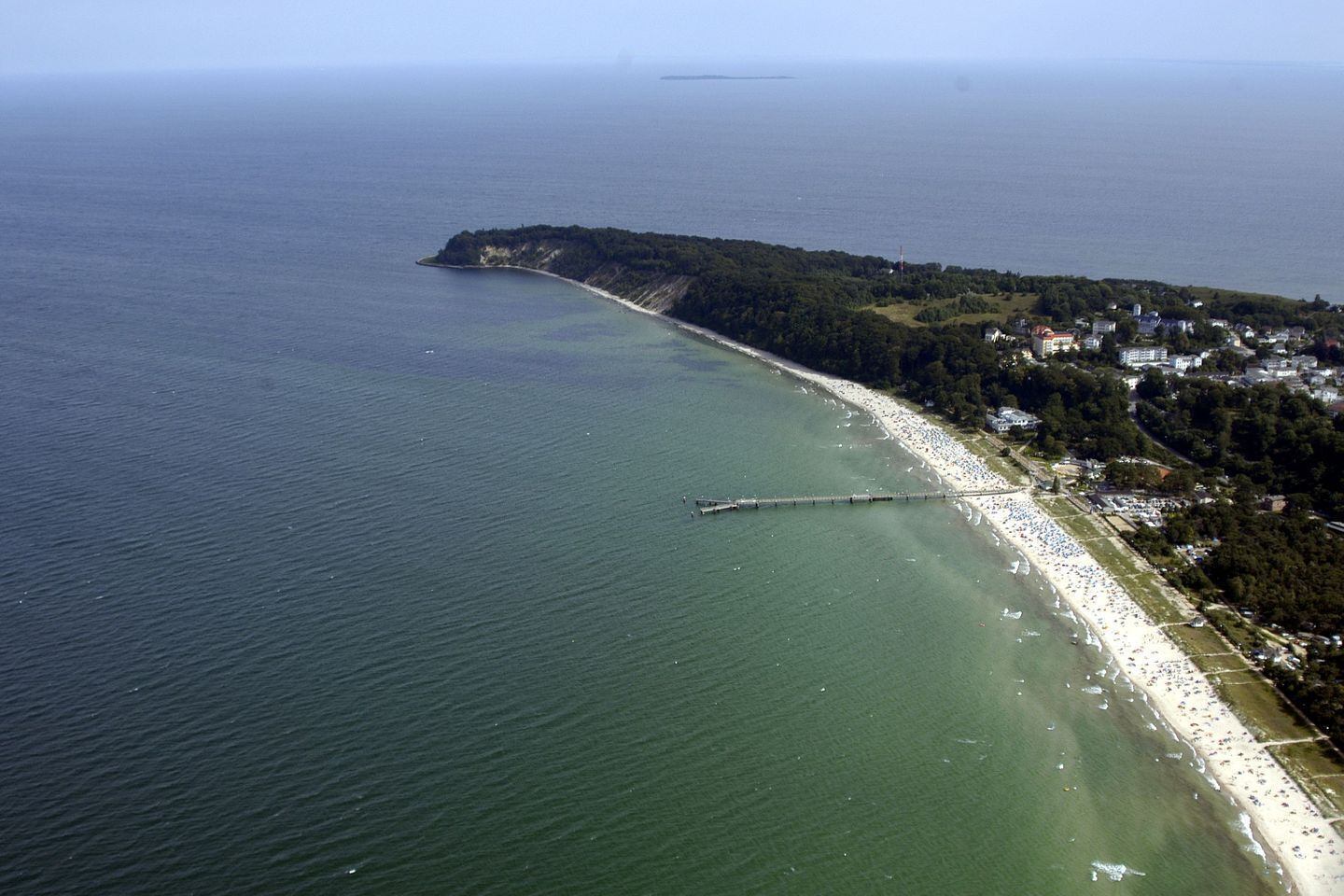 Oversigtsbillede af et kystområde med en lang mole, der strækker sig ud i havet, en sandstrand og en tæt bevokset halvø.