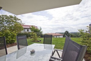 Un patio al aire libre con mesa y sillas de vidrio, rodeado de follaje y con vistas a casas al fondo.