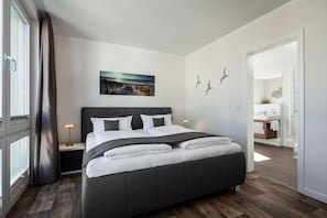 A modern bedroom with a large bed, a bedside table, a nightstand, a window with curtains, and a view of the bathroom.