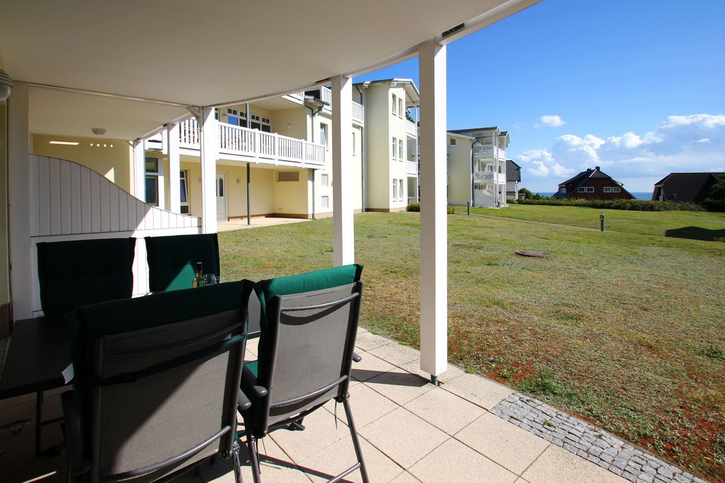 Eine überdachte Terrasse mit zwei Stühlen und einem Tisch, mit Blick auf eine Rasenfläche und ein Gebäude mit Balkonen.