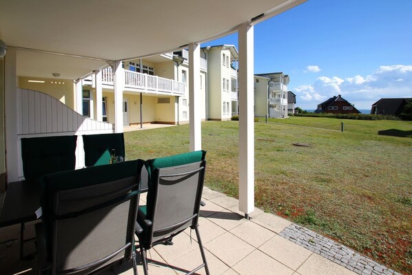 A covered patio with two chairs and a table, overlooking a grassy area and a building with balconies.