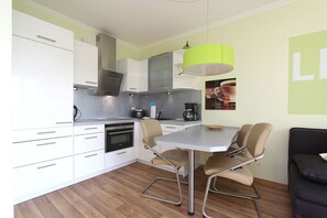 A compact kitchen with white cabinets, a built-in oven, a coffee maker, and a dining table with four chairs. - Patio, free use of the AHOI adventure pool and sauna - Apartment 31 with ocean view - (Göhren)
