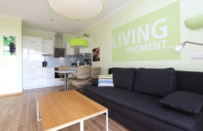 A modern living room with a sofa, a wooden table, and a kitchen in the background. - Terrace, free use of the AHOI adventure pool and sauna - Sea view apartment 31 - (Göhren)