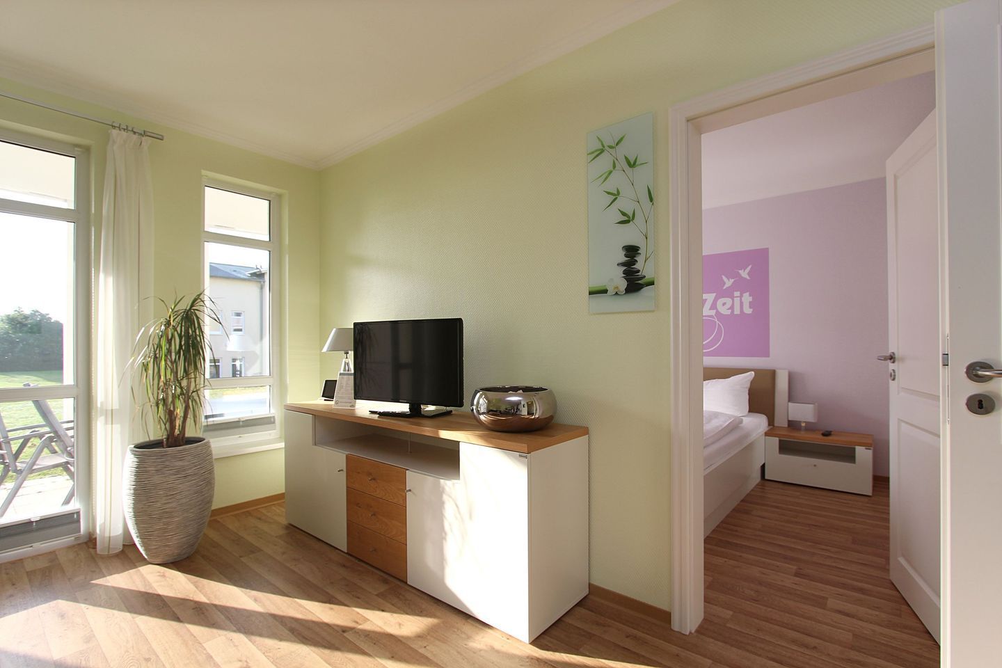 A hotel room with a wooden desk, a flat-screen TV, a potted plant, and a bed with a nightstand.
