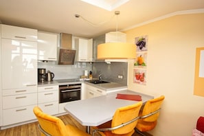 A modern kitchen with white cabinets, a built-in oven, a coffee maker, and a sink. There are yellow chairs around a table and a large yellow pendant light above the sink.