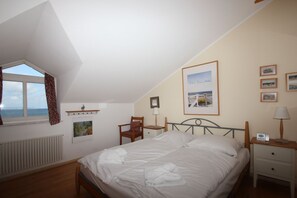 Ein Schlafzimmer mit Bett, Sessel, Nachttisch, Lampe, Bilderrahmen und einem Fenster mit Vorhängen.