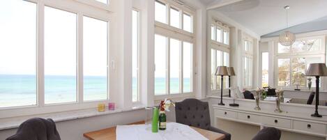 Una habitación con un ventanal grande con vista al mar, una mesa de madera con mantel blanco y una estantería con libros.