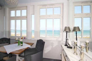 A room with large windows overlooking a beach, a dining table with chairs, and a small cabinet with decorative items.