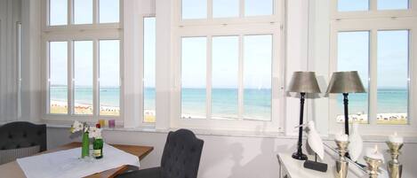 Ein Zimmer mit großen Fenstern und Blick auf einen Strand, einem Esstisch mit Stühlen und einer kleinen Vitrine mit Dekoration.