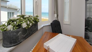 Una habitación con vista a la playa, una maceta negra en el alféizar de la ventana, una mesa de madera con una caja blanca y una silla negra.