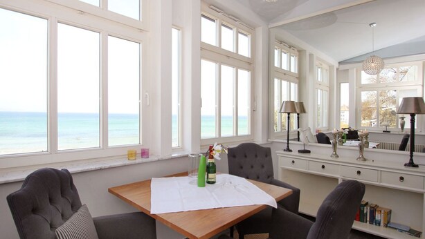 Una habitación con un ventanal grande con vista al mar, una mesa de madera con mantel blanco y una estantería con libros.
